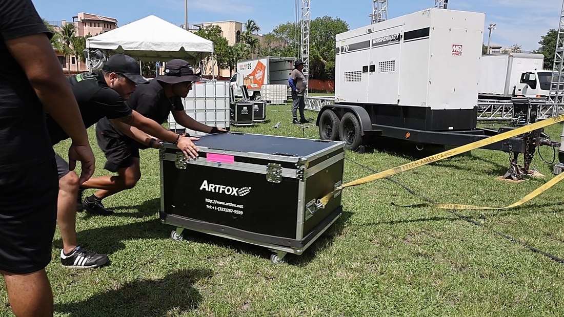 ATM crew moving a road case at the Peacock Park build, with the towable generator and water ballast tanks behind